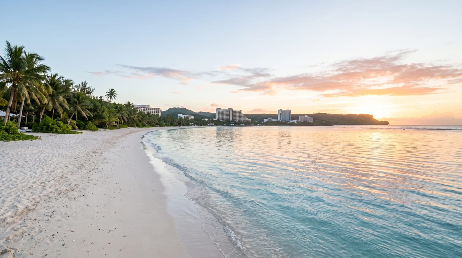 Weißer Sandstrand der Tumon Bay auf Guam bei Sonnenaufgang, türkisfarbenes Wasser, Palmen und Hotels in der Ferne