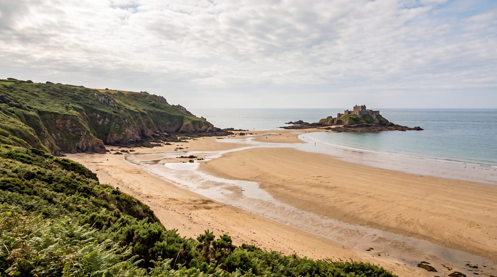 Bucht auf Jersey bei Ebbe mit breitem Sandstrand, grünen Klippen und einer Burg auf einem Inselchen