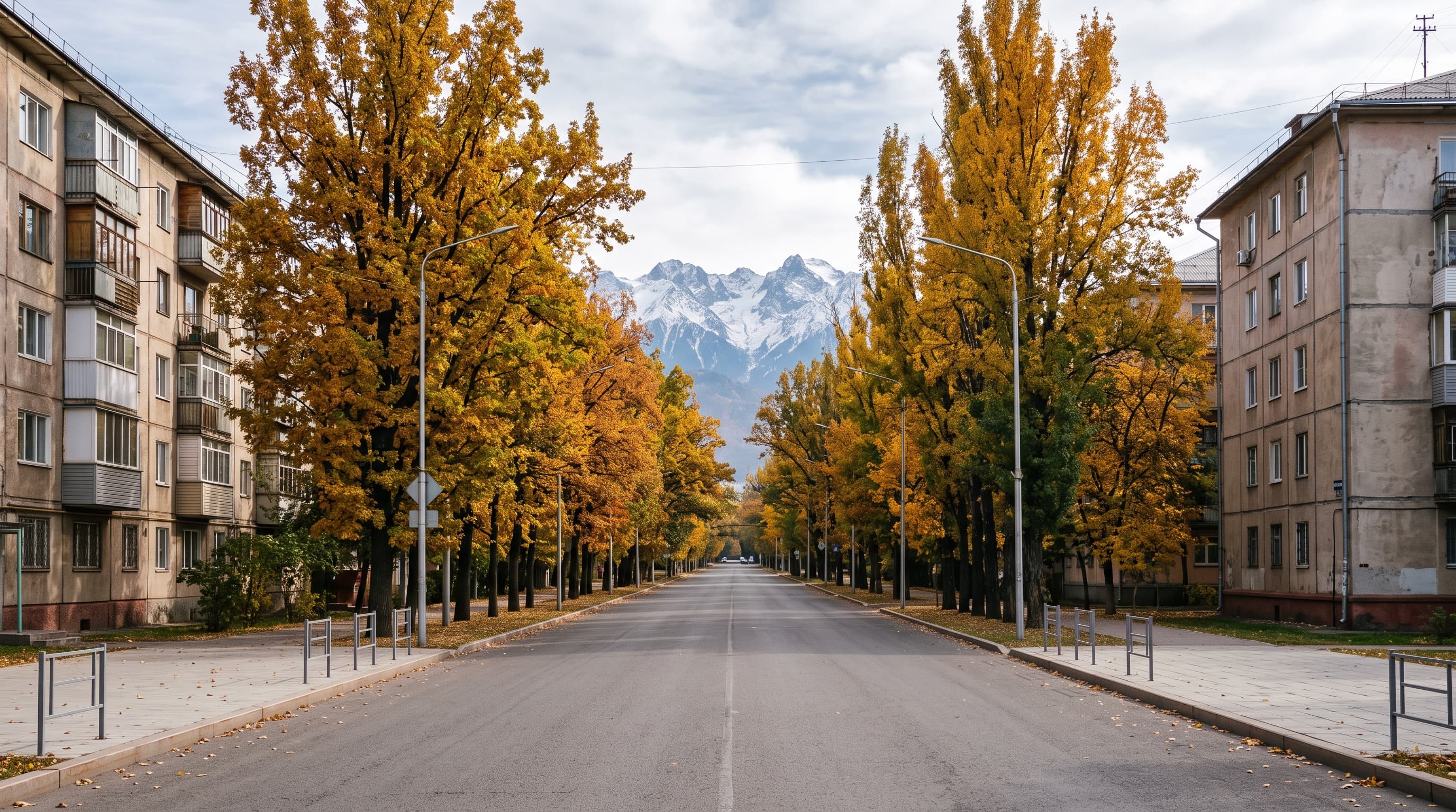 Rue résidentielle calme d'Almaty bordée de grands arbres à l'ombre dense, immeubles bas d'époque soviétique, silhouette des montagnes du Tian Chan en arrière-plan