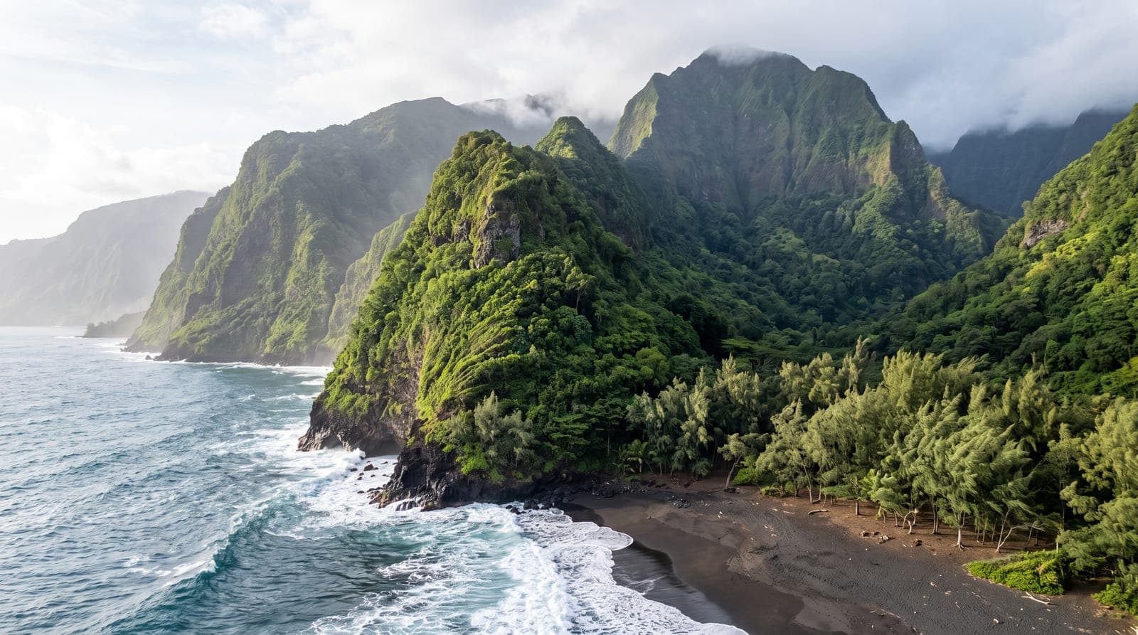 Küste von La Réunion mit grünen tropischen Klippen, schwarzem Sandstrand und Gipfeln im Dunst